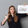 O femeie care ține o sticlă de apă arată spre un panou cu „Detox Suplimente” pe un fundal gri.